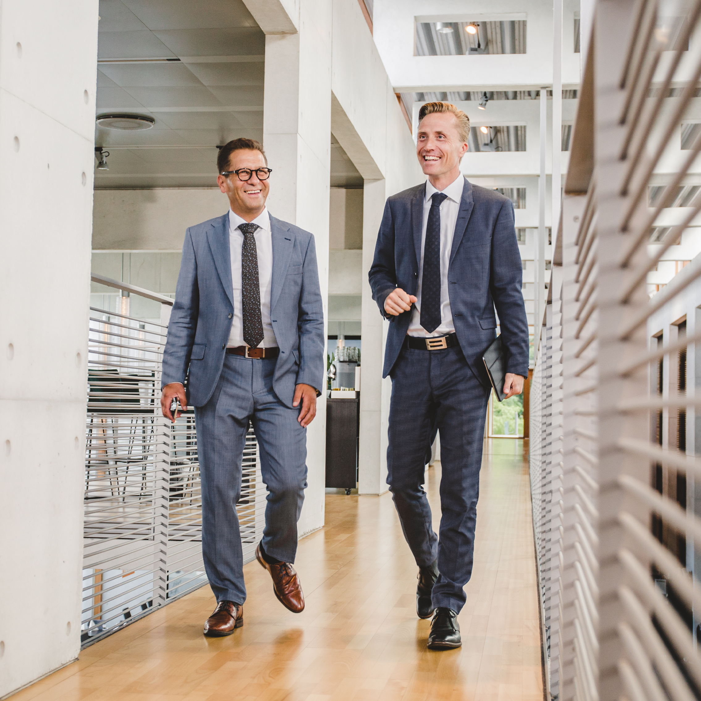 Two professionally dressed men in business suits walking and smiling in a modern office building, representing a positive and professional work environment.